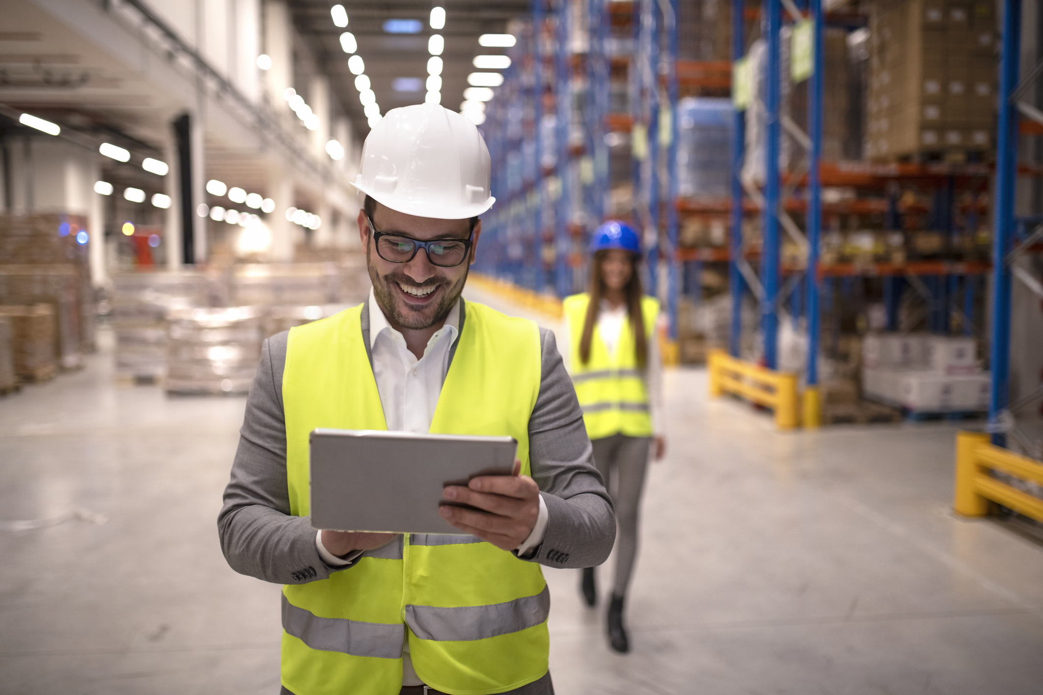Warehouse manager reading report on tablet about successful delivery and distribution in warehouse logistics center. In background coworker checking inventory and productivity in storage area.