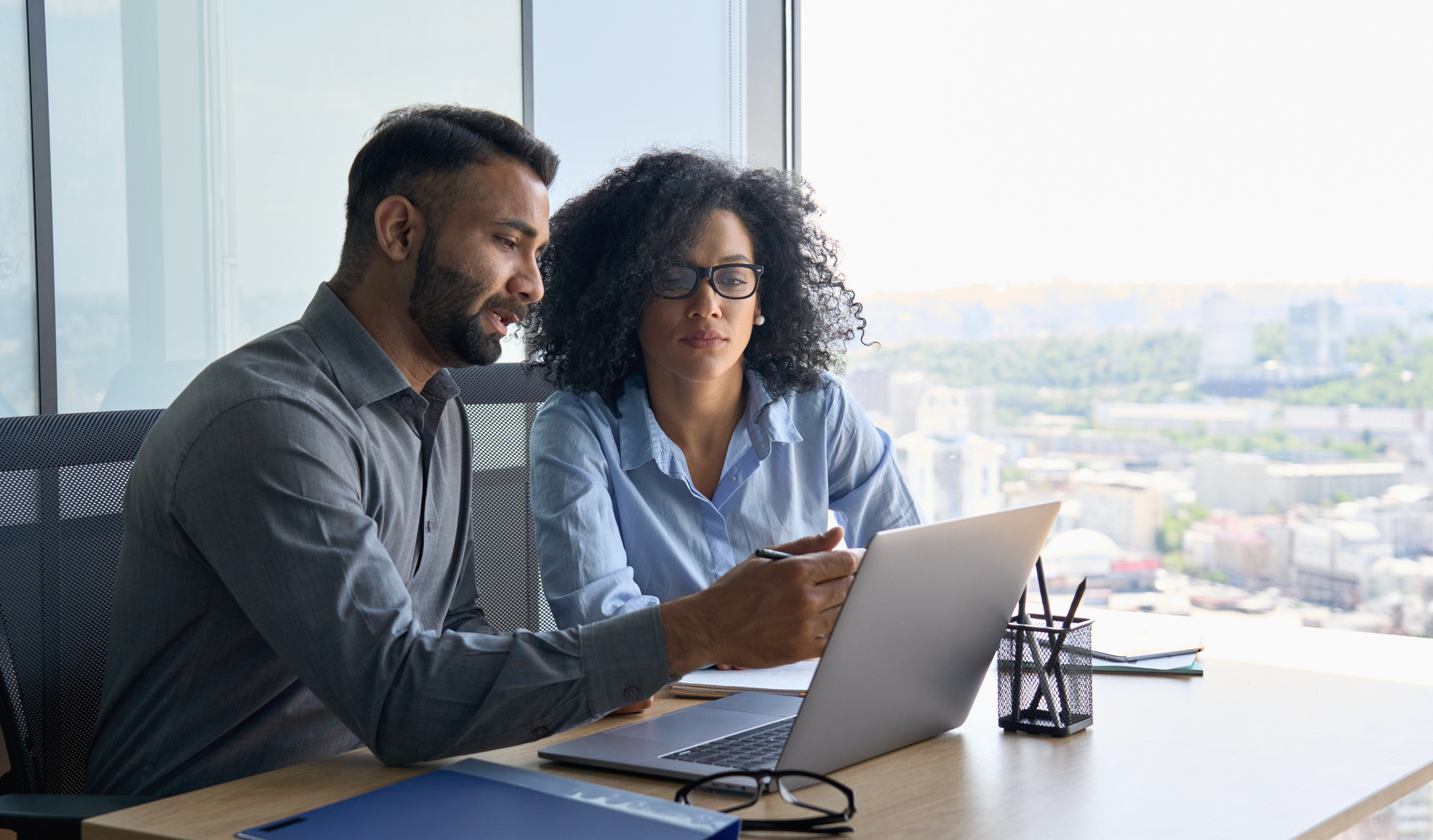 Multiethnic colleagues sitting at desk looking at laptop computer in office. Indian male ceo executive manager mentor giving consultation on financial operations to female African American colleague intern using laptop sitting in modern office near panoramic window.
